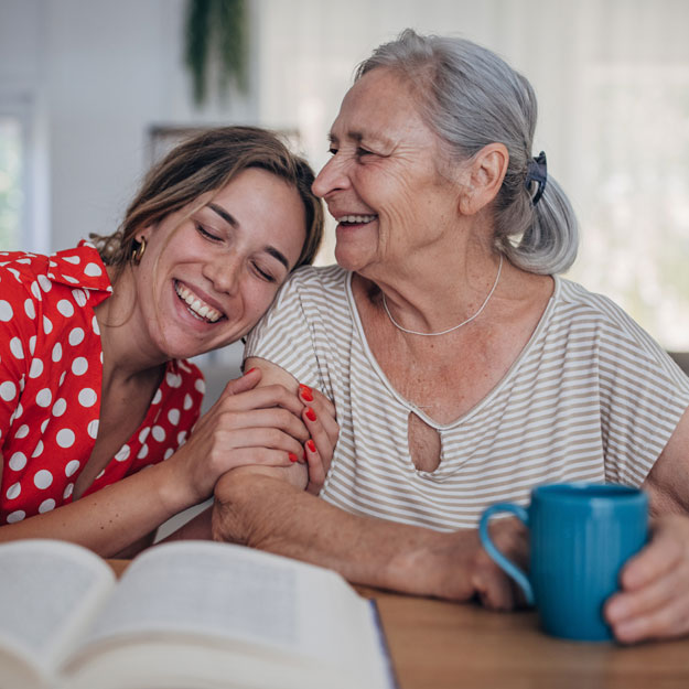 Two women who seem familiar with each other. The elder has her hand on a turquoise coffee mug, though the contents aren't seen. The younger woman is wearing a red and white top, smiling as she leans on the other woman's shoulder with a book open in front of them.