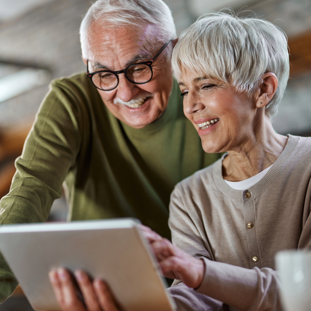 Woman holds a tablet while a man looks over her shoulder. They are both smiling at something on screen.